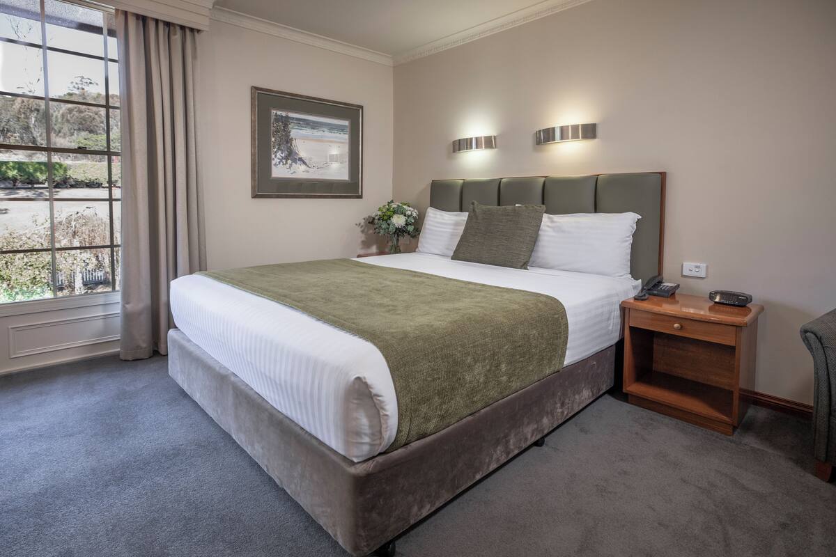 A neatly arranged hotel room with a large bed, a bedside table with a lamp, and a window with curtains.