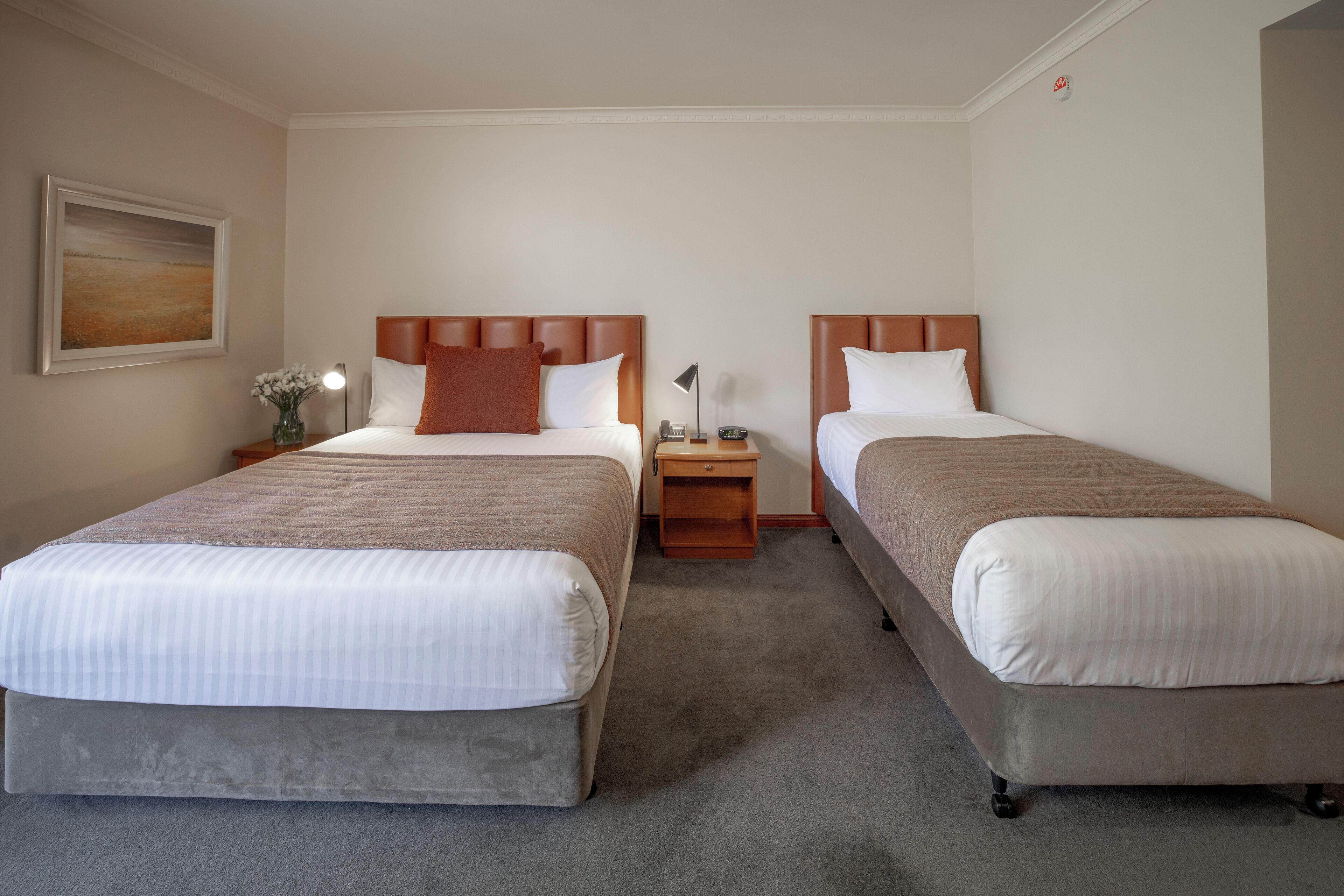 Habitación de hotel con dos camas, una mesita de noche con lámpara, un jarrón con flores y un cuadro en la pared.