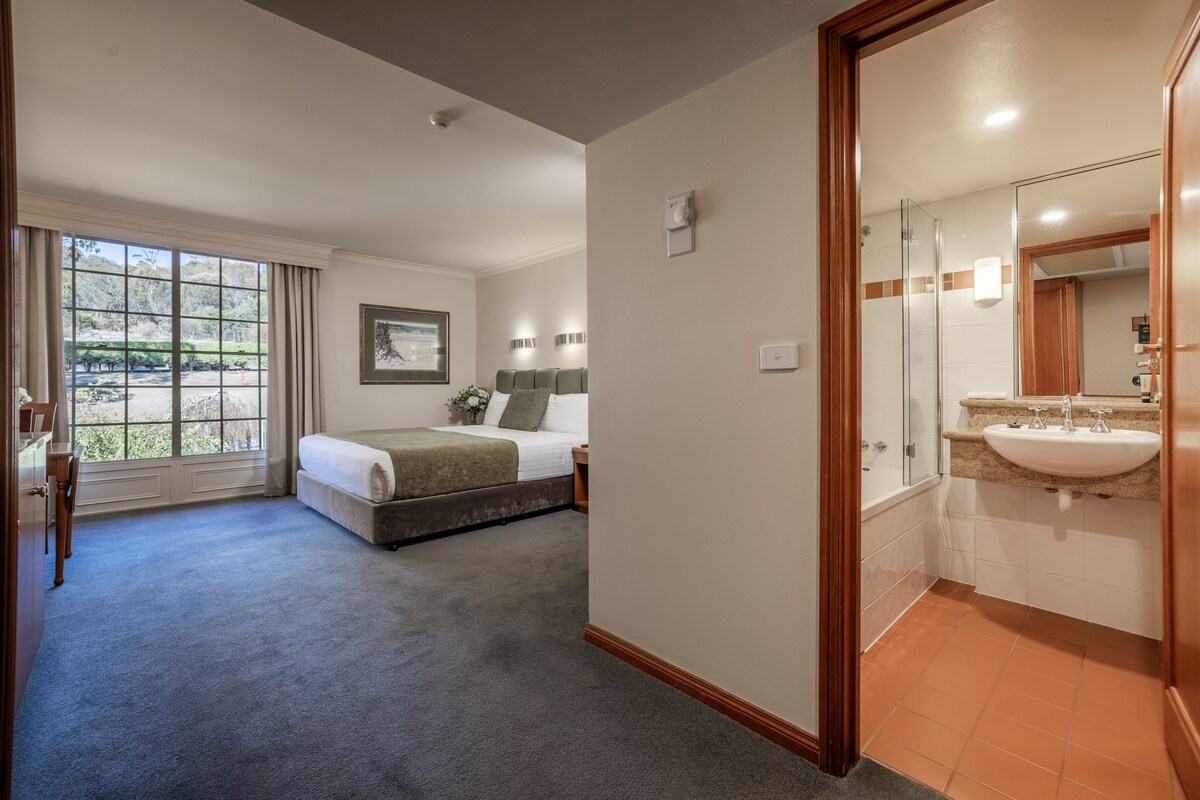 A hotel room with a large bed, a desk, a chair, a window with curtains, a bathroom with a sink and mirror, and a framed picture on the wall.