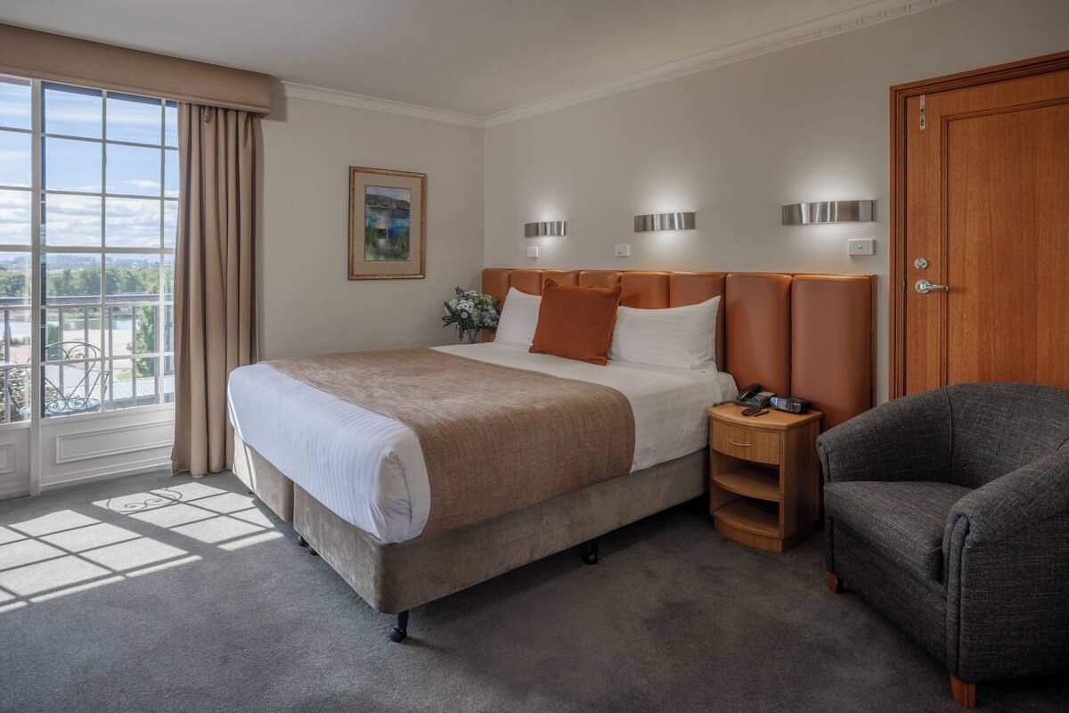 A hotel room with a large bed, a nightstand with a phone, a chair, and a view of a cityscape through the window.