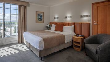 Habitación de hotel con una cama grande, una mesita de noche con un teléfono, una silla y vista a un paisaje urbano a través de la ventana.