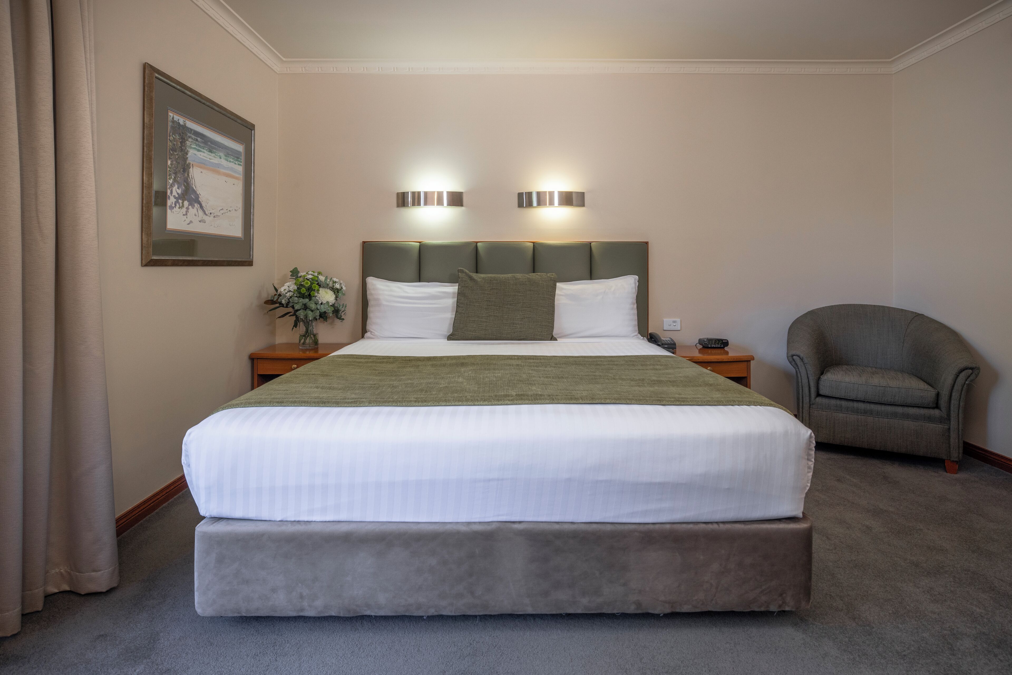 Un letto ben rifatto con una coperta verde, due abat-jour, una sedia e un quadro appeso al muro.