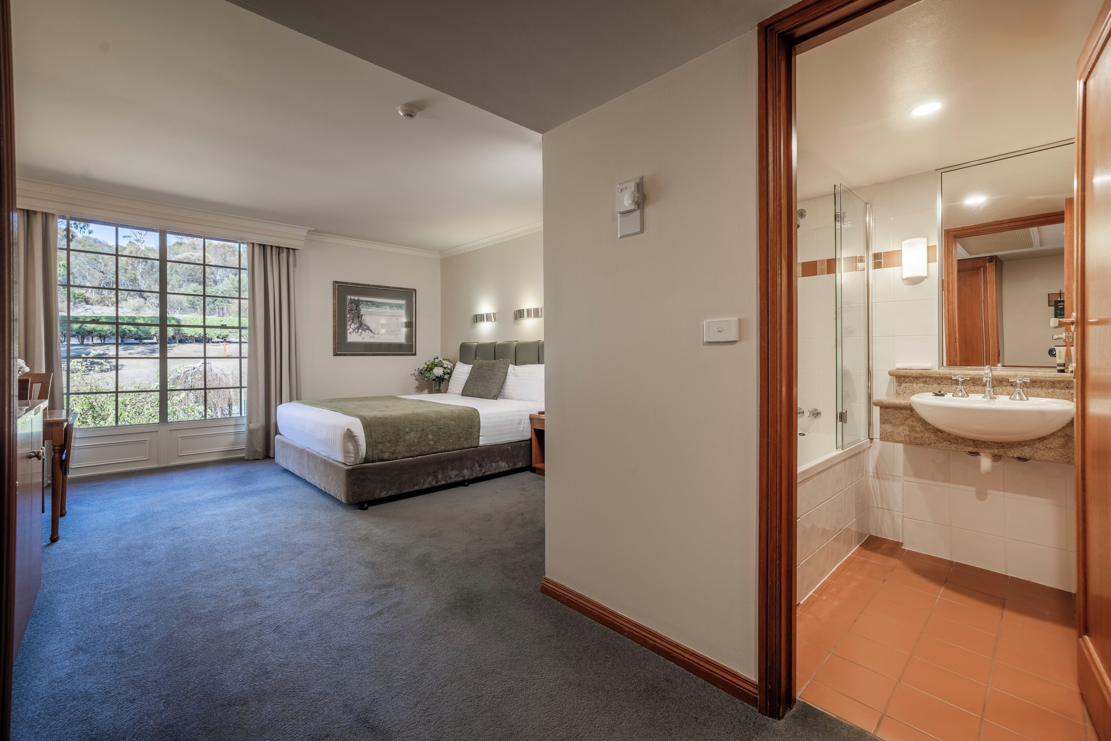 A hotel room with a large window, a bed, a bedside table, a mirror, and a bathroom with a sink and toilet.