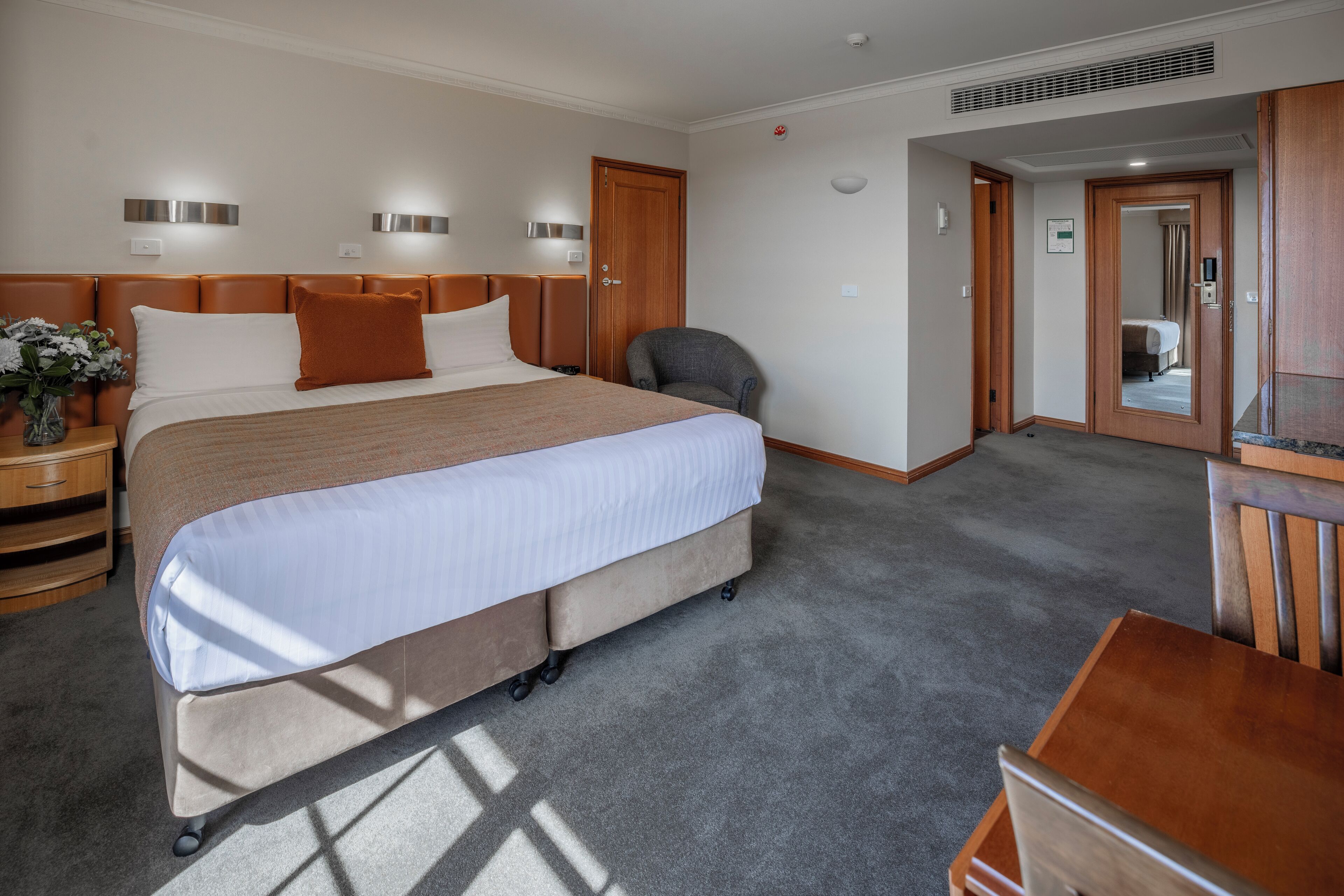 Habitación de hotel con una cama grande, un escritorio y una silla. Hay una mesita de noche con un jarrón de flores y una lámpara. El piso está alfombrado y la puerta es de madera.