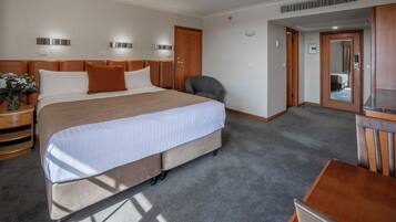 A hotel room with a large bed, a desk, and a chair. There is a nightstand with a vase of flowers and a lamp. The room has a carpeted floor and a wooden door.
