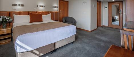 Habitación de hotel con una cama grande, un escritorio y una silla. Hay una mesita de noche con un jarrón de flores y una lámpara. El piso está alfombrado y la puerta es de madera.