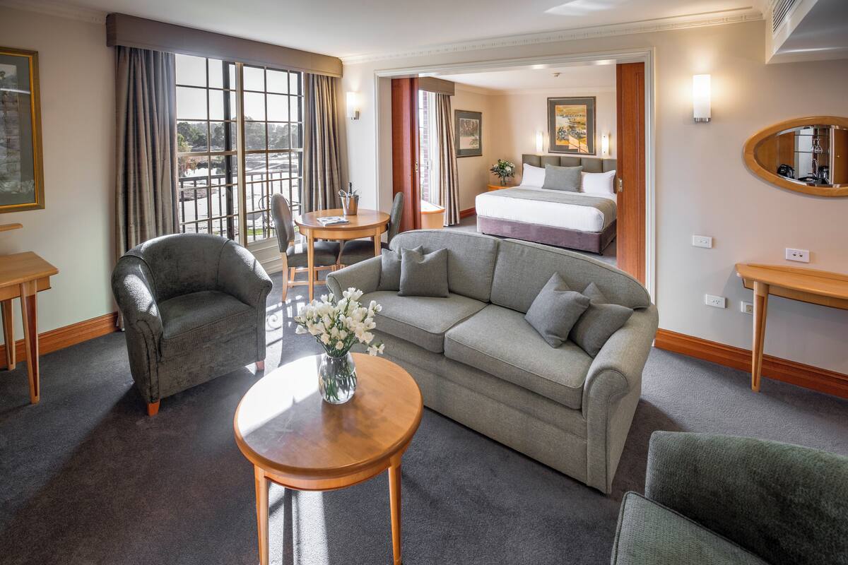 A hotel room with a sofa, a chair, a small table with a vase of flowers, a bed, and a large window with curtains.