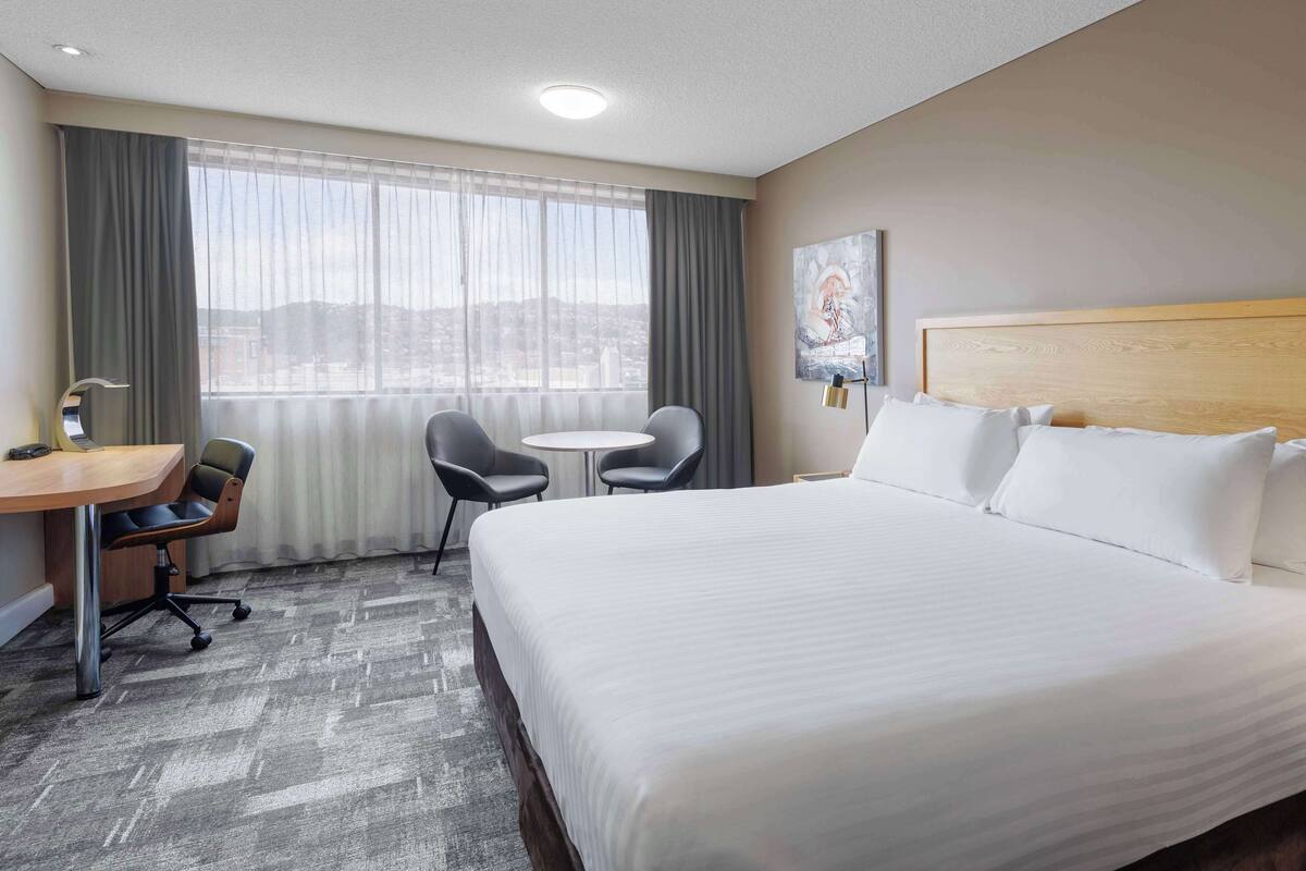 A hotel room with a large bed, a desk, two chairs, a small table, and a window with curtains.