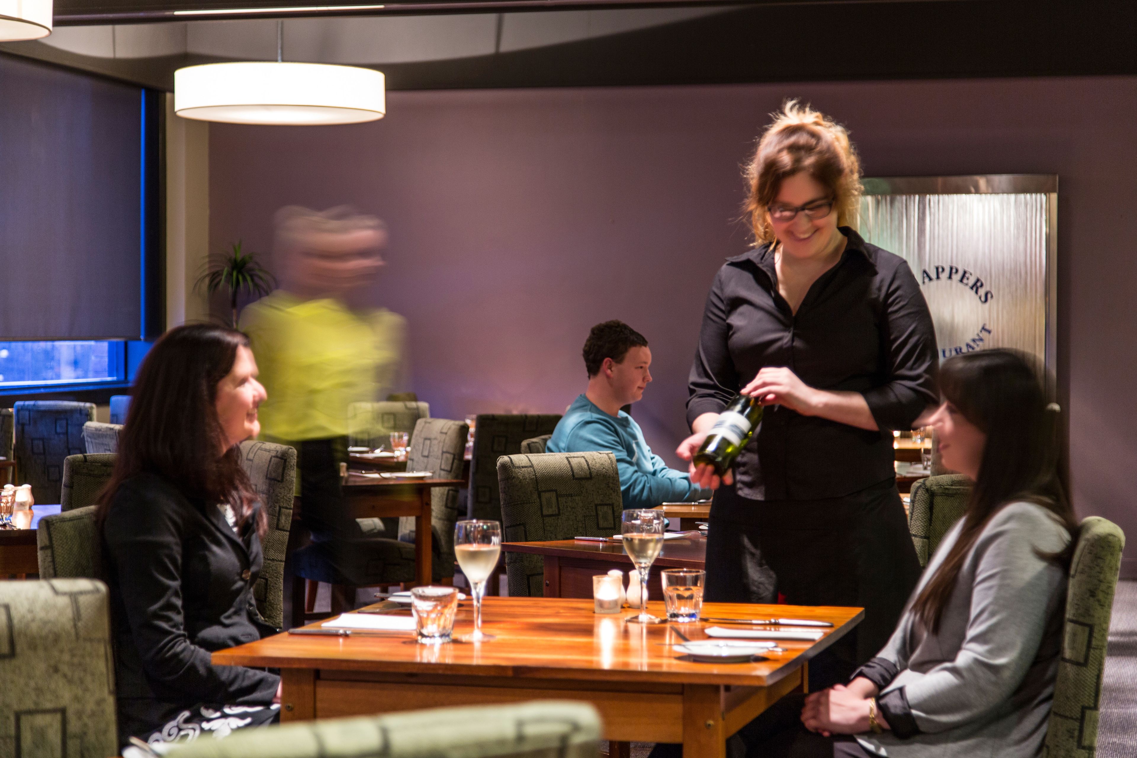 A restaurant setting with a waiter pouring a drink, tables with chairs, and a sign in the background.