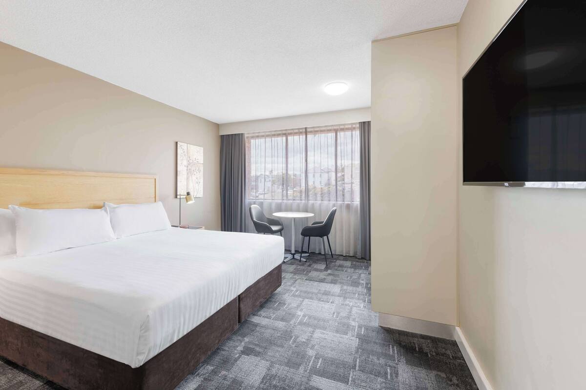A hotel room with a large bed, a small table with two chairs, a TV mounted on the wall, and a window with curtains.