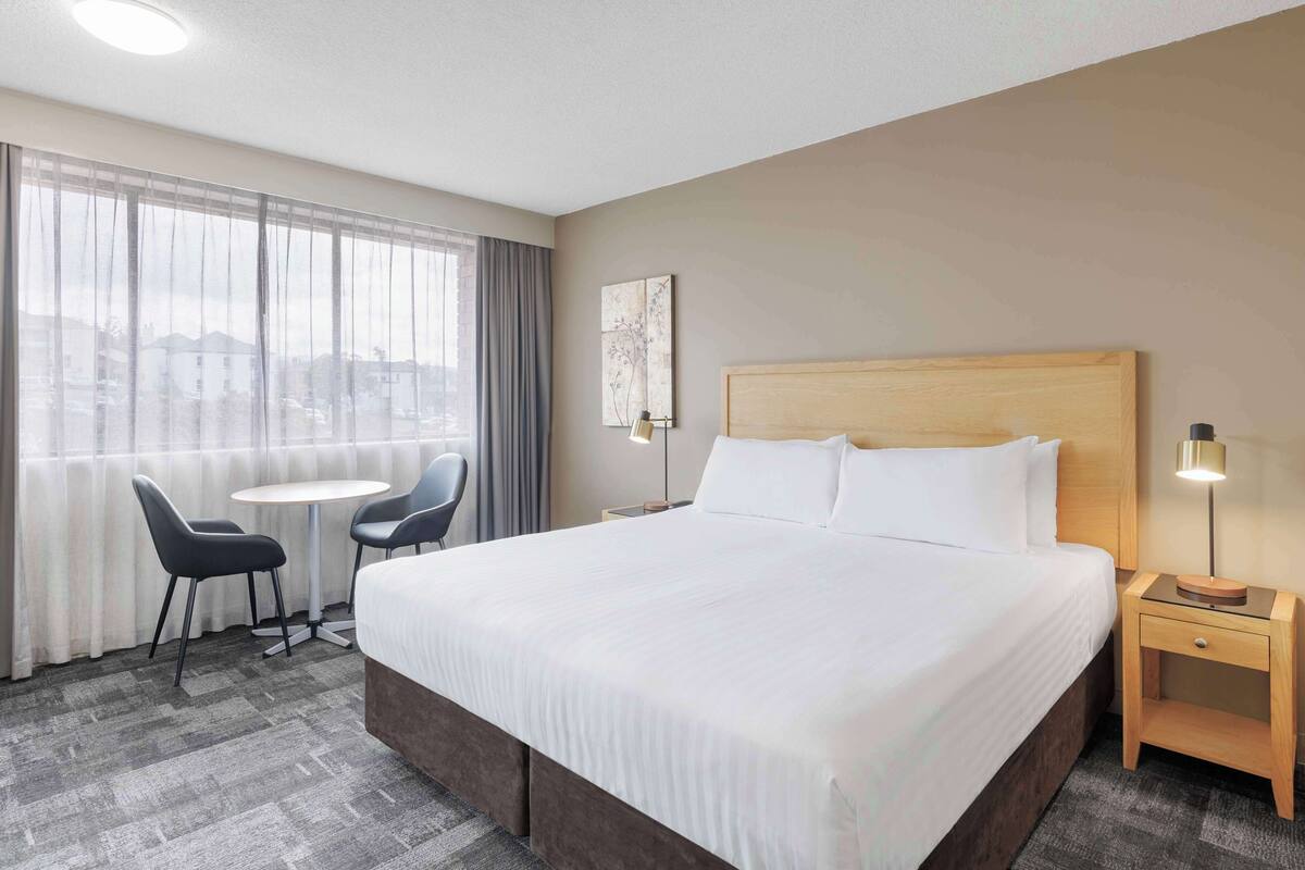 A hotel room with a large bed, a small table, two chairs, a nightstand with a lamp, and a window with curtains.