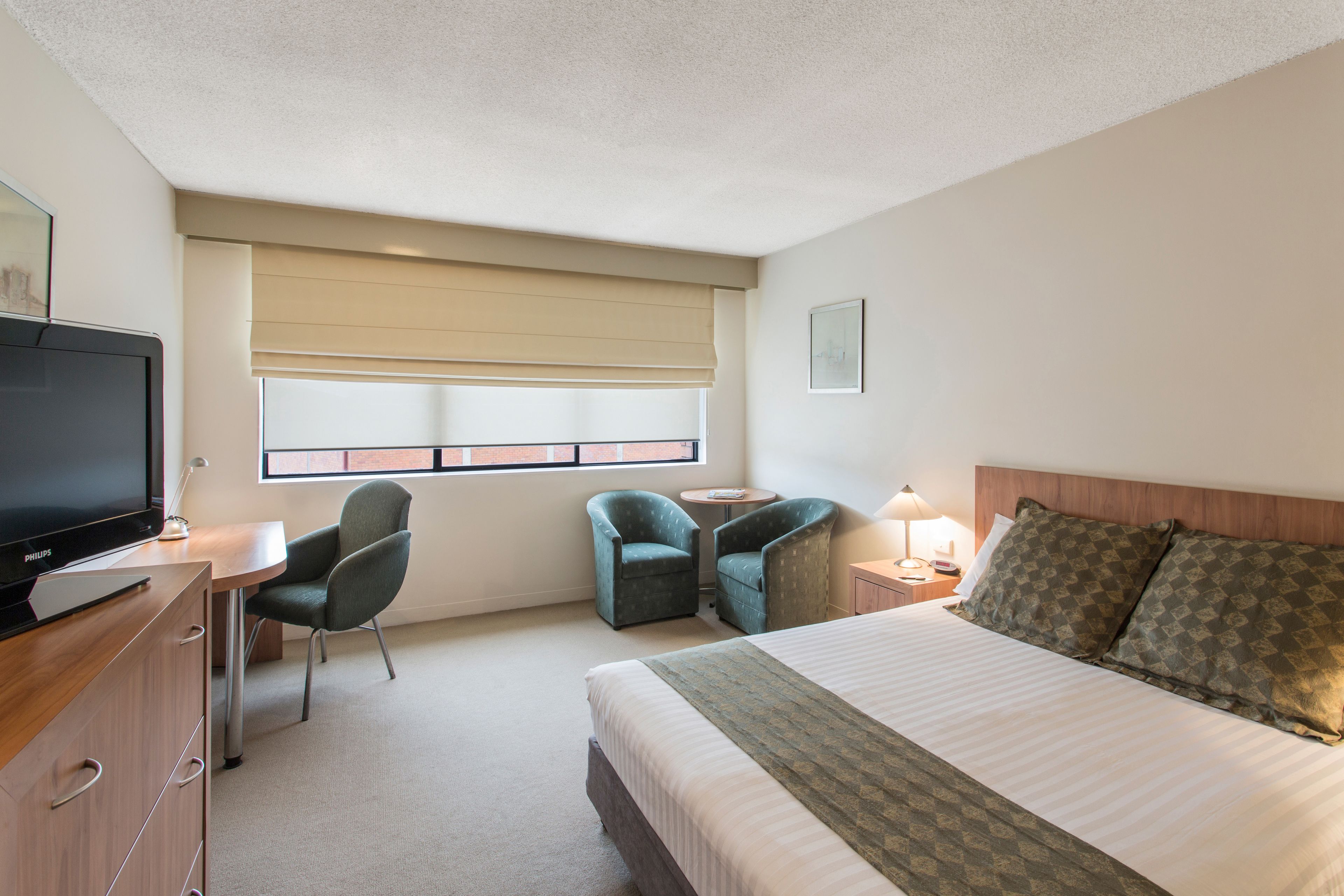 A hotel room with a bed, two armchairs, a desk with a television, a window with blinds, and a bedside table with a lamp.