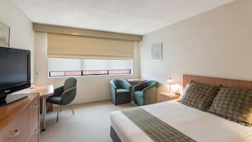 A hotel room with a bed, two armchairs, a desk with a television, a window with blinds, and a bedside table with a lamp.