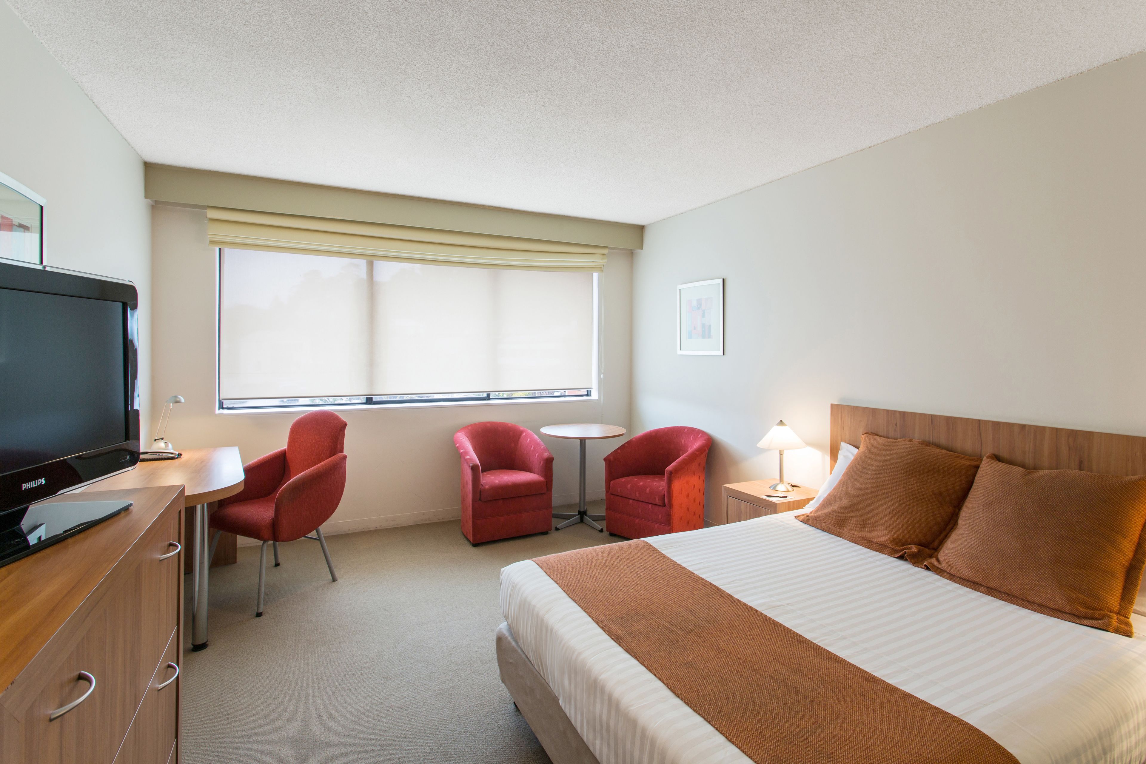 Une chambre d’hôtel avec un lit, un bureau avec une télévision, deux chaises rouges, une petite table et une fenêtre avec des stores.