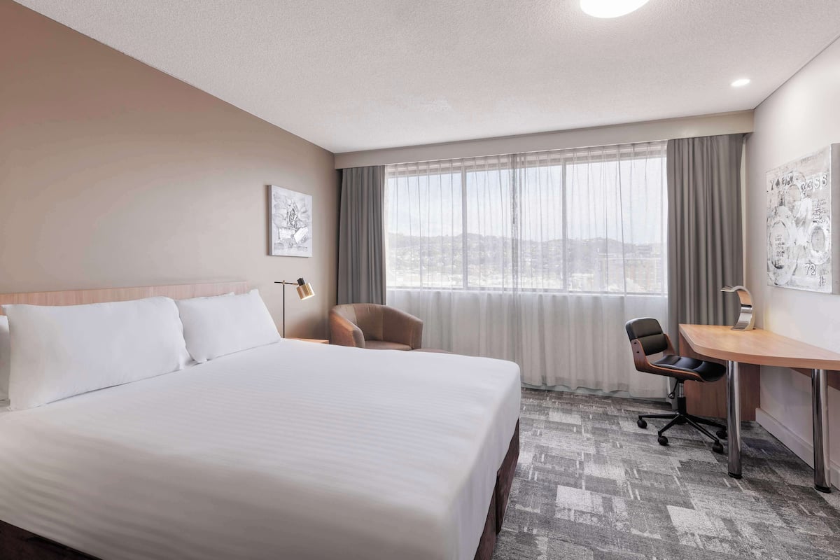 A hotel room with a large bed, a desk, a chair, and a view of the city through the window.