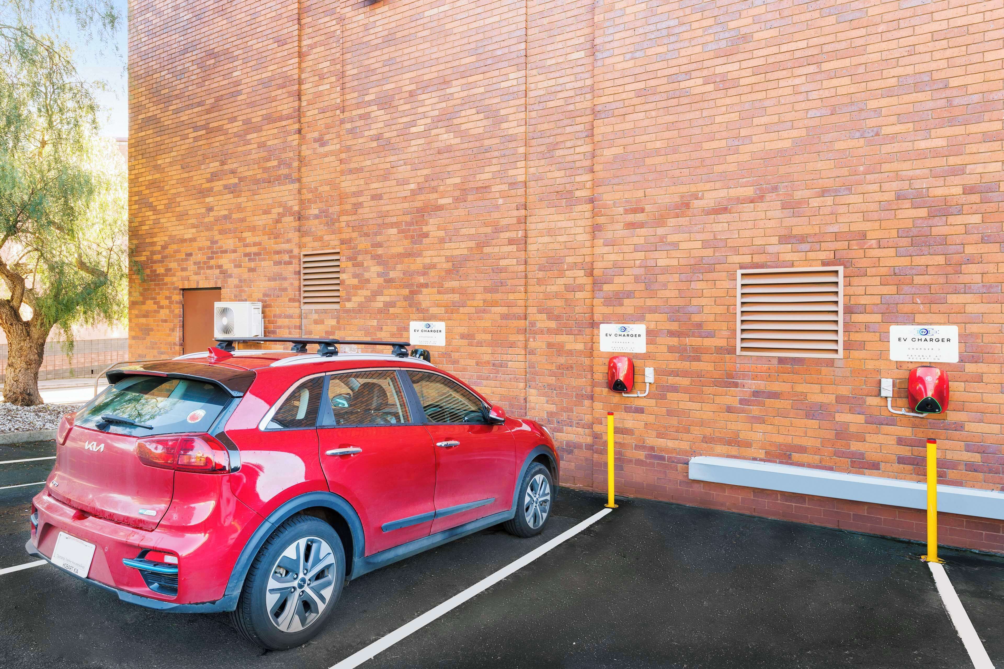 A red car parked in a designated EV charging station area with a brick building in the background.