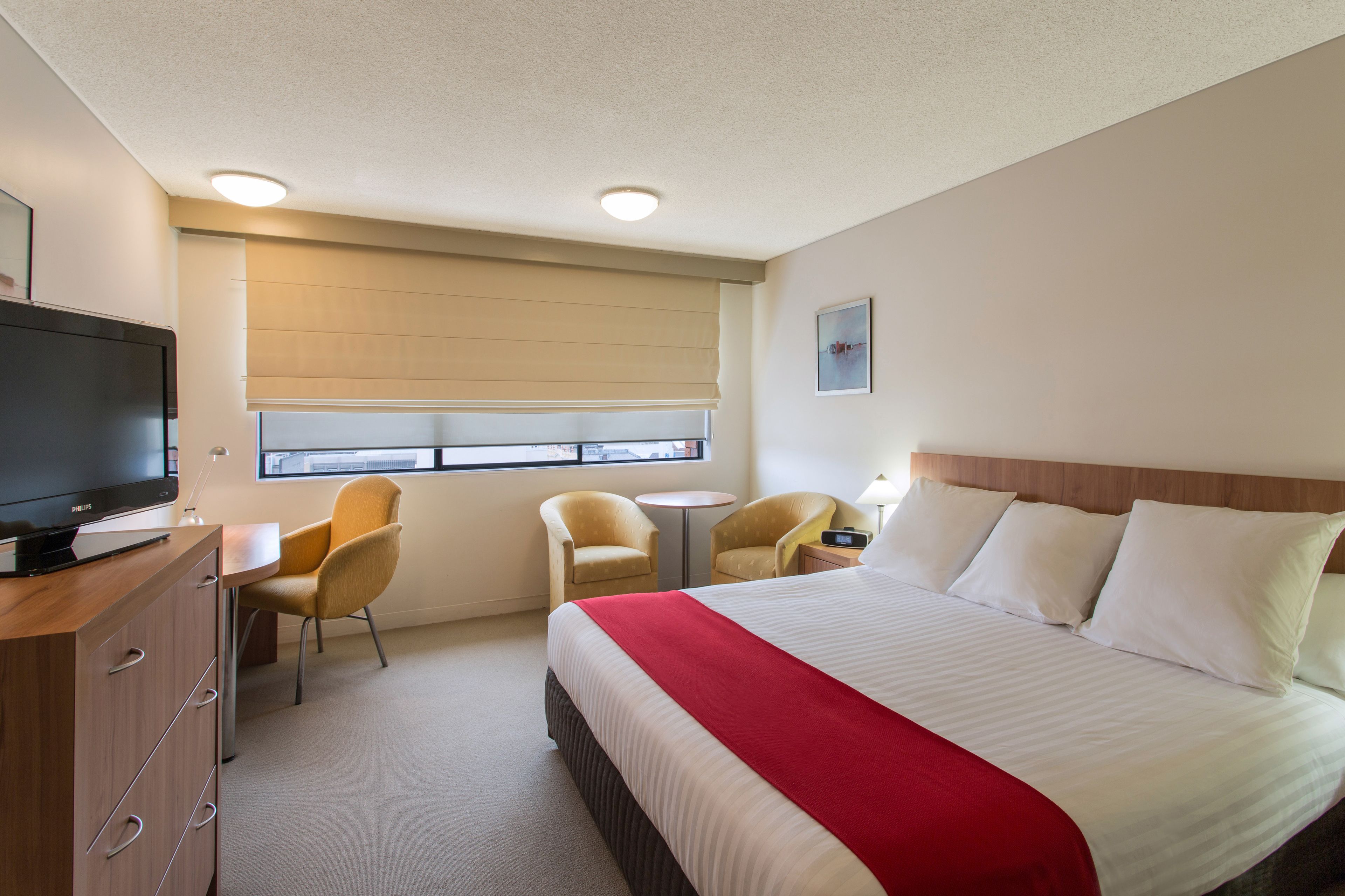 Une chambre d’hôtel avec un lit, une télévision, deux chaises, une petite table et une fenêtre avec des stores.