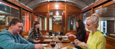 A dining car with wooden paneling and a curved ceiling, featuring a table set for four people with wine glasses and plates.