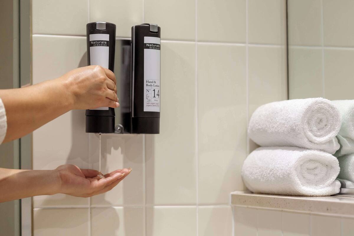 A hand dispensing lotion from a dispenser labeled "Naturals Remedies" in a bathroom with white tiles and a stack of white towels.