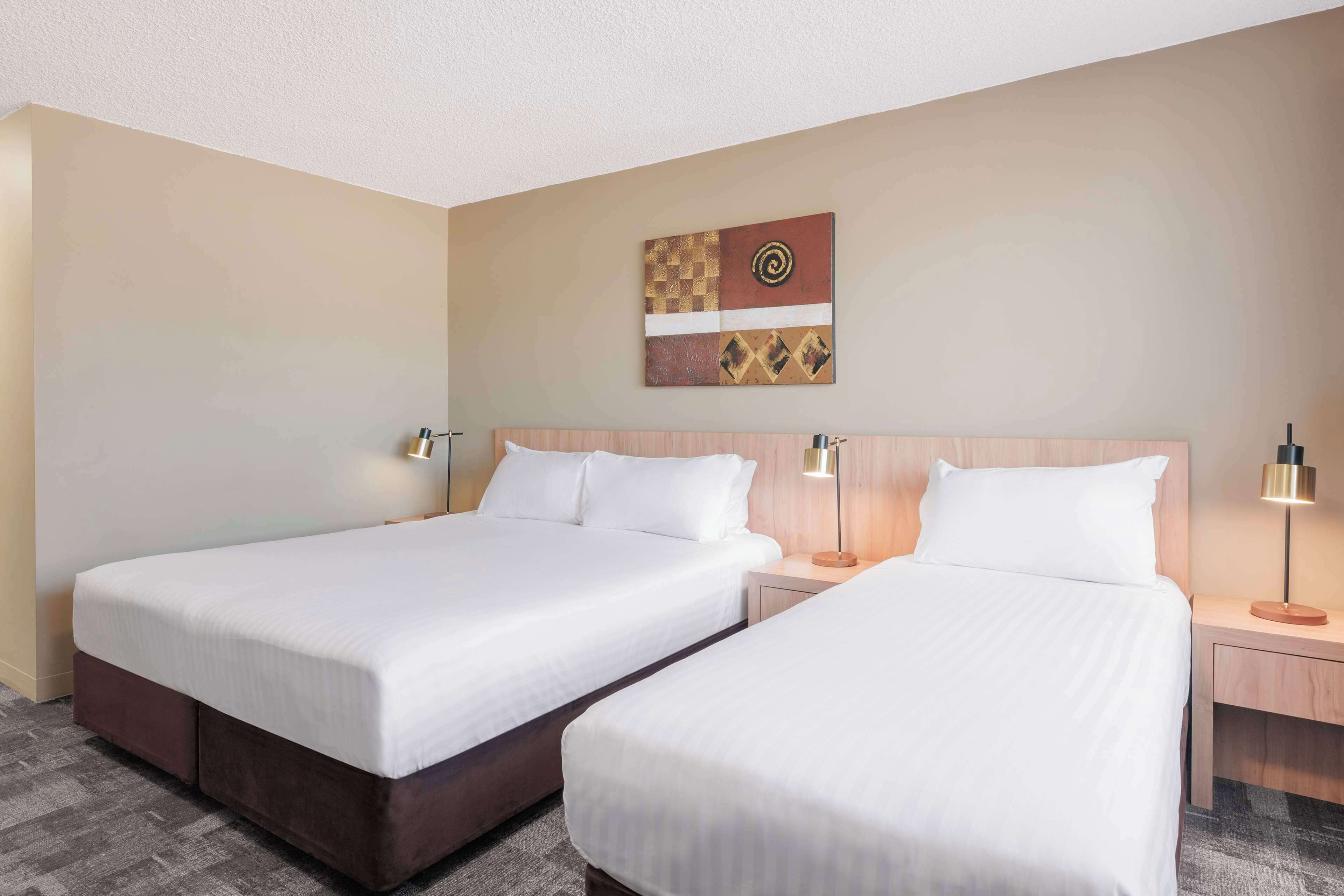 Habitación de hotel con dos camas individuales, cabeceros de madera, mesitas de noche con lámparas y una obra de arte colgada en la pared.