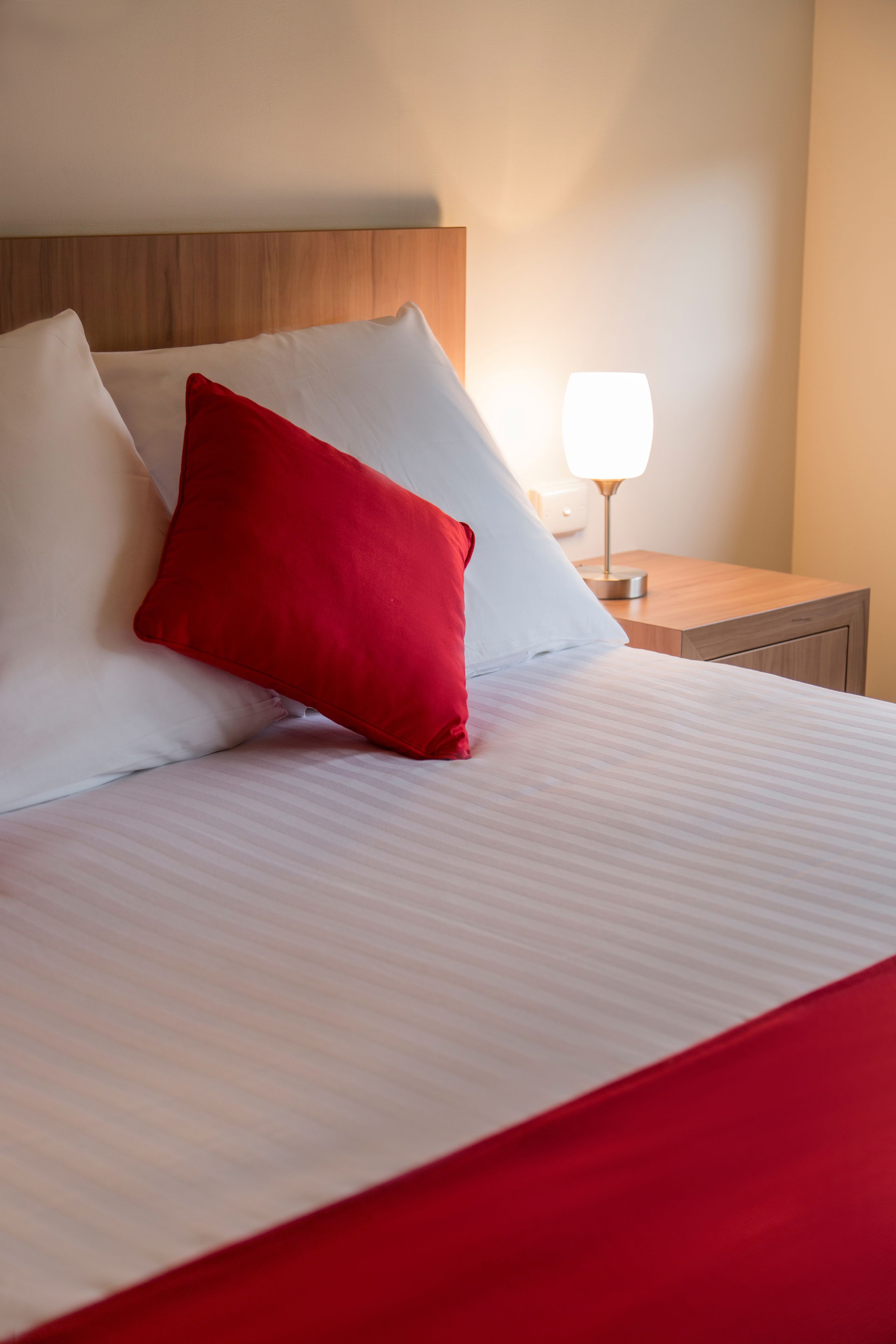 Una cama bien hecha con almohadas blancas y rojas, un cabecero de madera y una mesita de noche con lámpara.