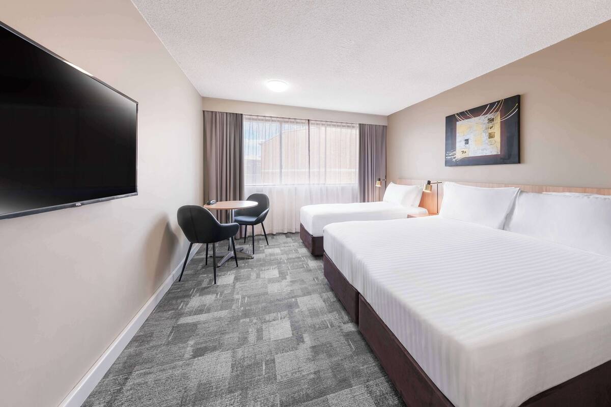 A hotel room with a flat-screen TV, two beds, a desk with chairs, and a large window with curtains.