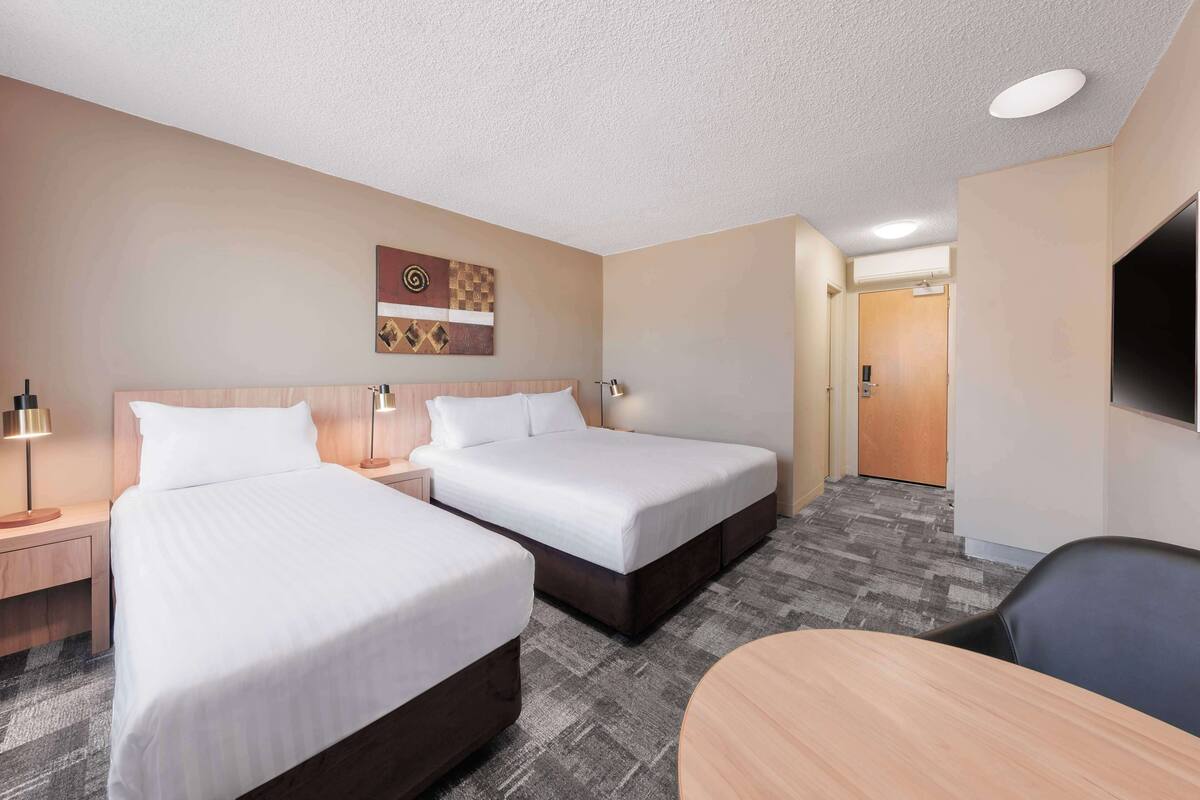 A hotel room with two beds, a desk, a chair, a TV, and a door leading to another room.