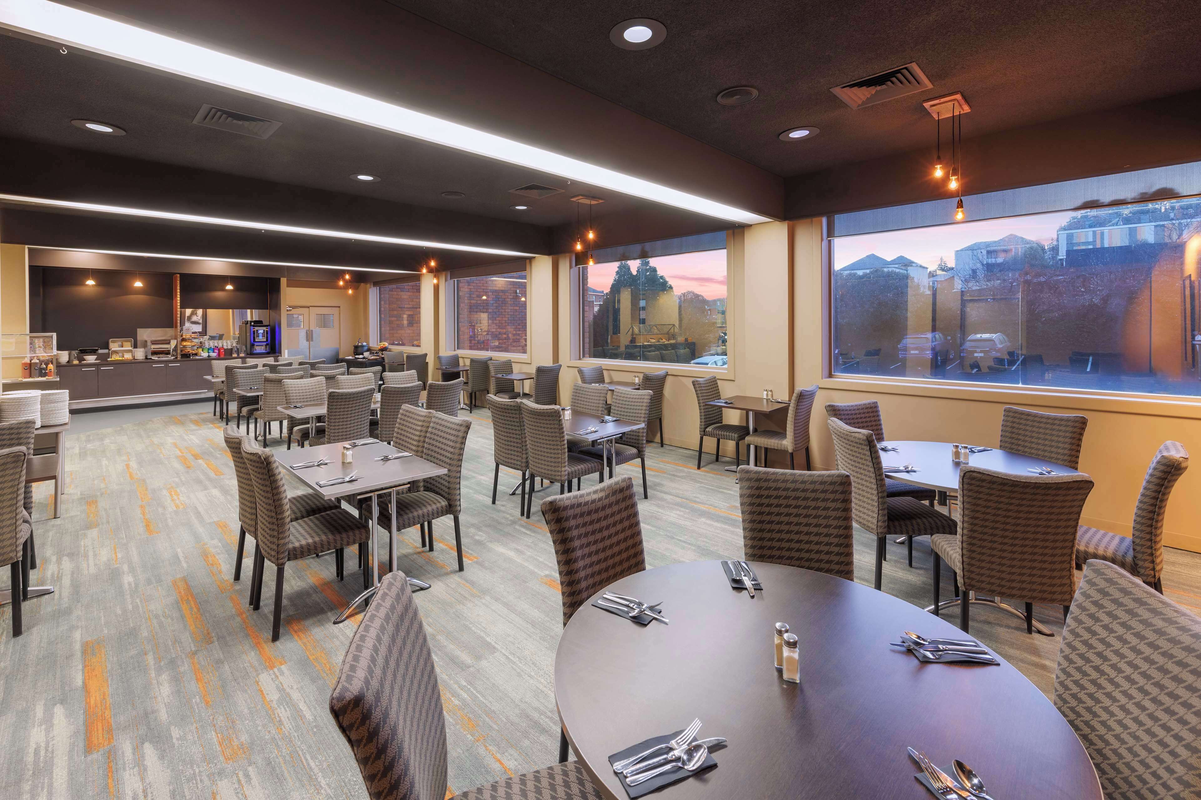 A hotel dining area with multiple tables and chairs, large windows with a view of the city, and a modern ceiling design.