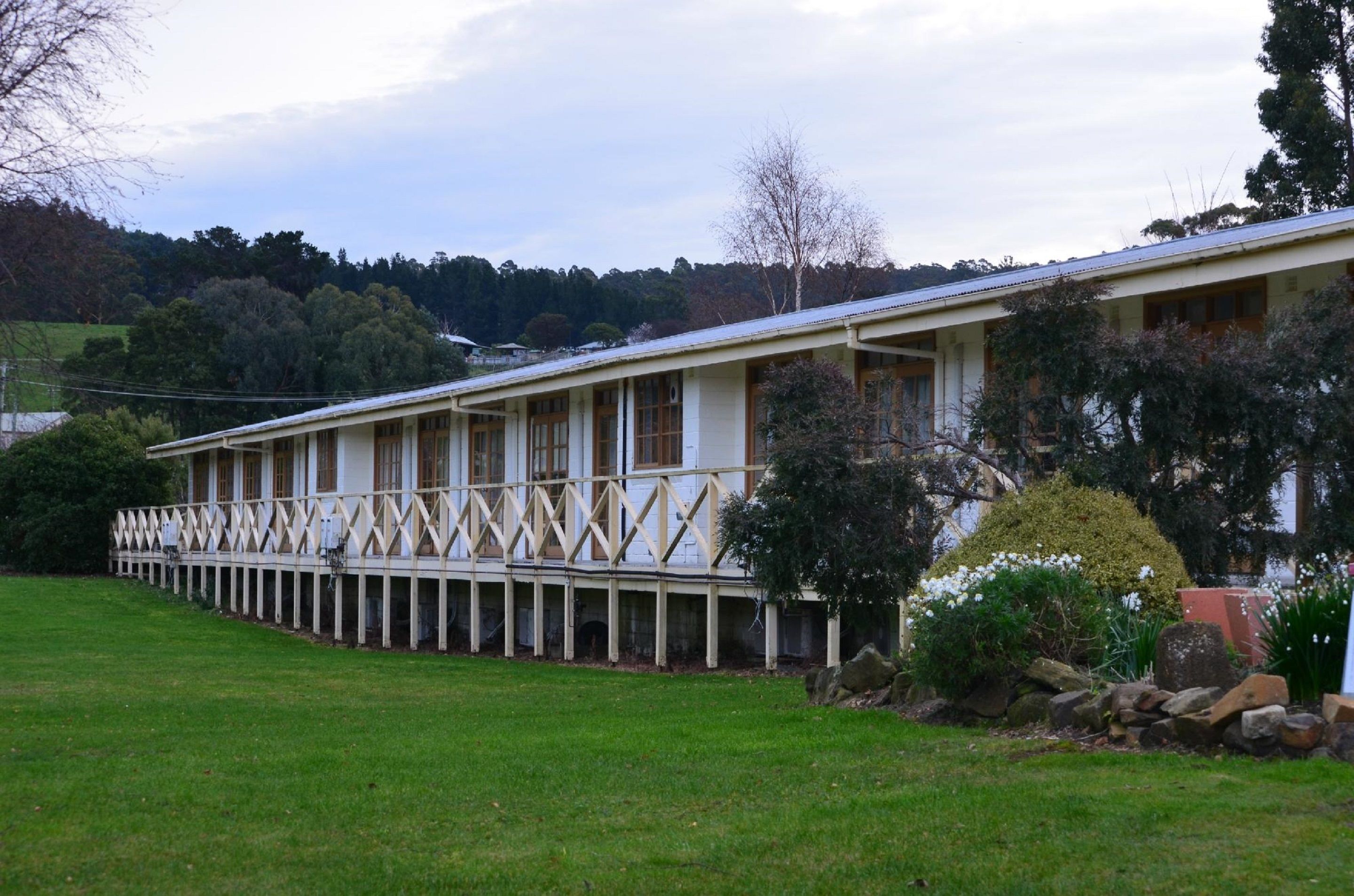 A long, single-story building with a white exterior and a flat roof. It features a series of windows along the side and a covered porch area.