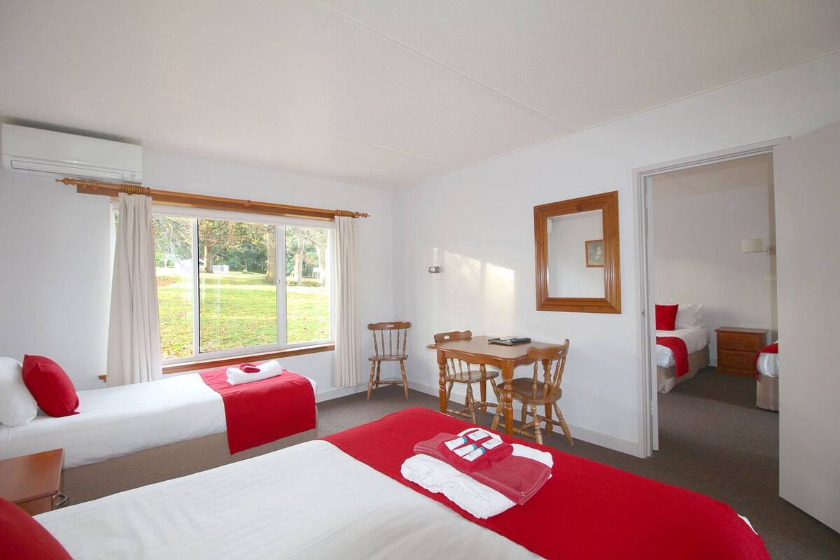 A hotel room with two beds, a dining table, chairs, a mirror, and a view of the outdoors.