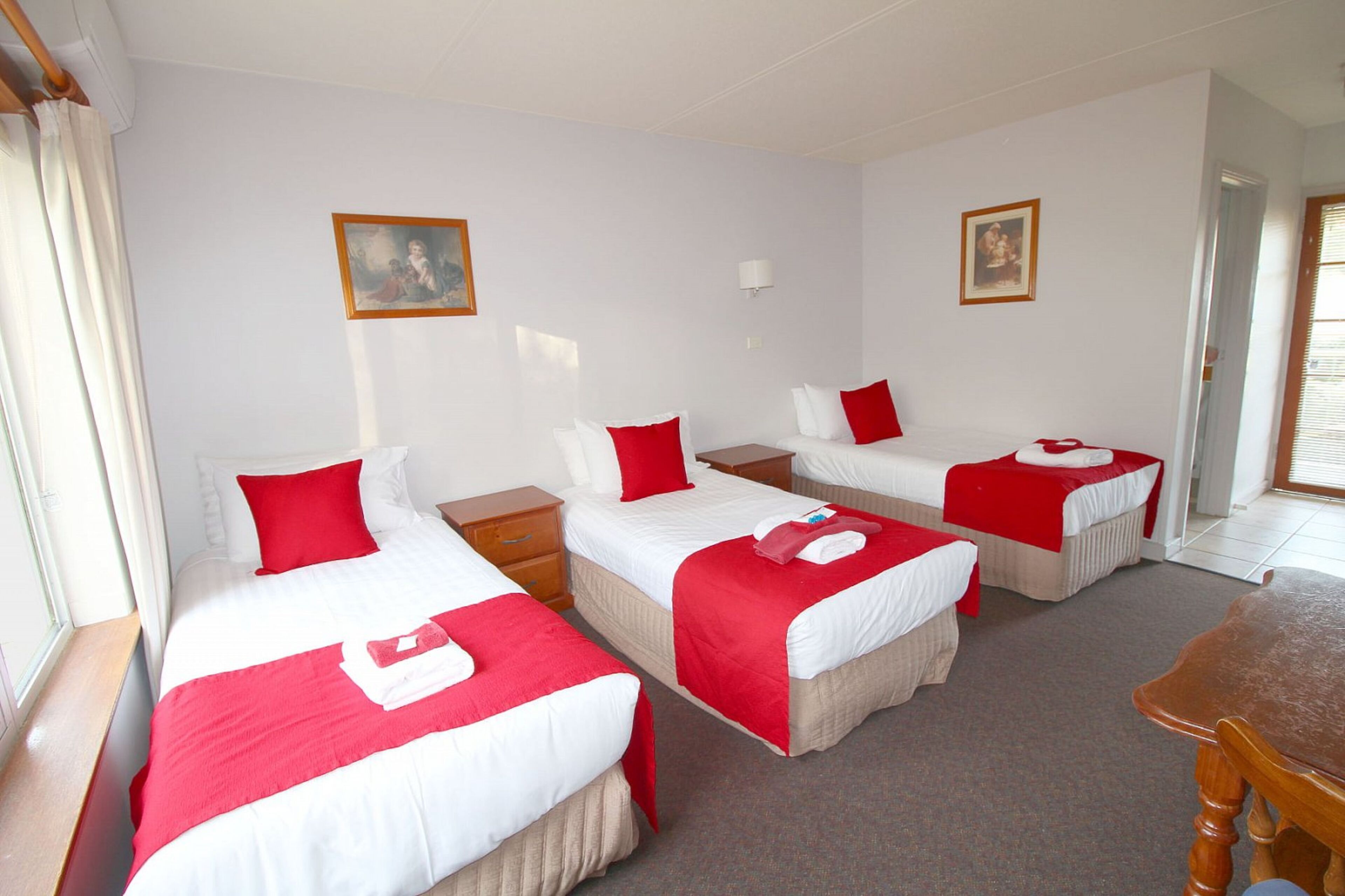 A hotel room with three single beds, each with a red and white bedspread and pillows. There are wooden nightstands, a small wooden table, and a window with curtains.