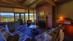 Un salon confortable avec des canapés à motifs, une cheminée et une table. De grandes fenêtres offrent une vue panoramique sur le paysage.