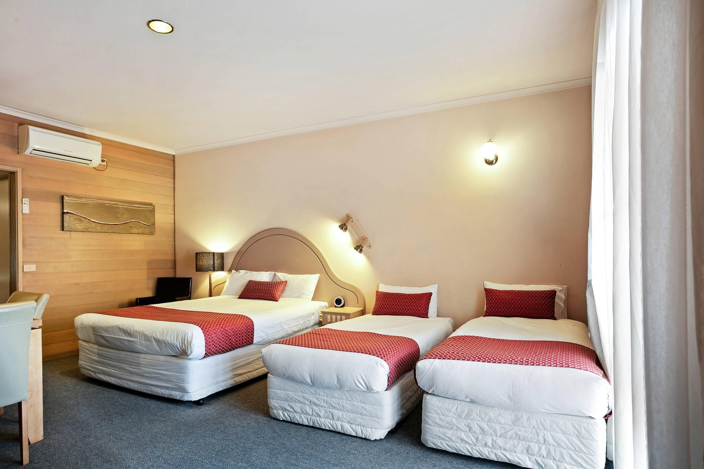 Quarto de hotel com três camas, uma escrivaninha e ar condicionado fixado na parede.