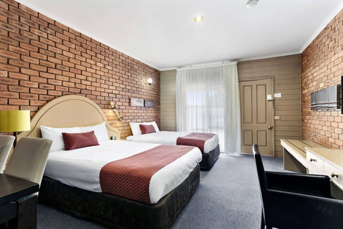 A hotel room with two beds, a desk, a chair, a TV, and a brick wall.