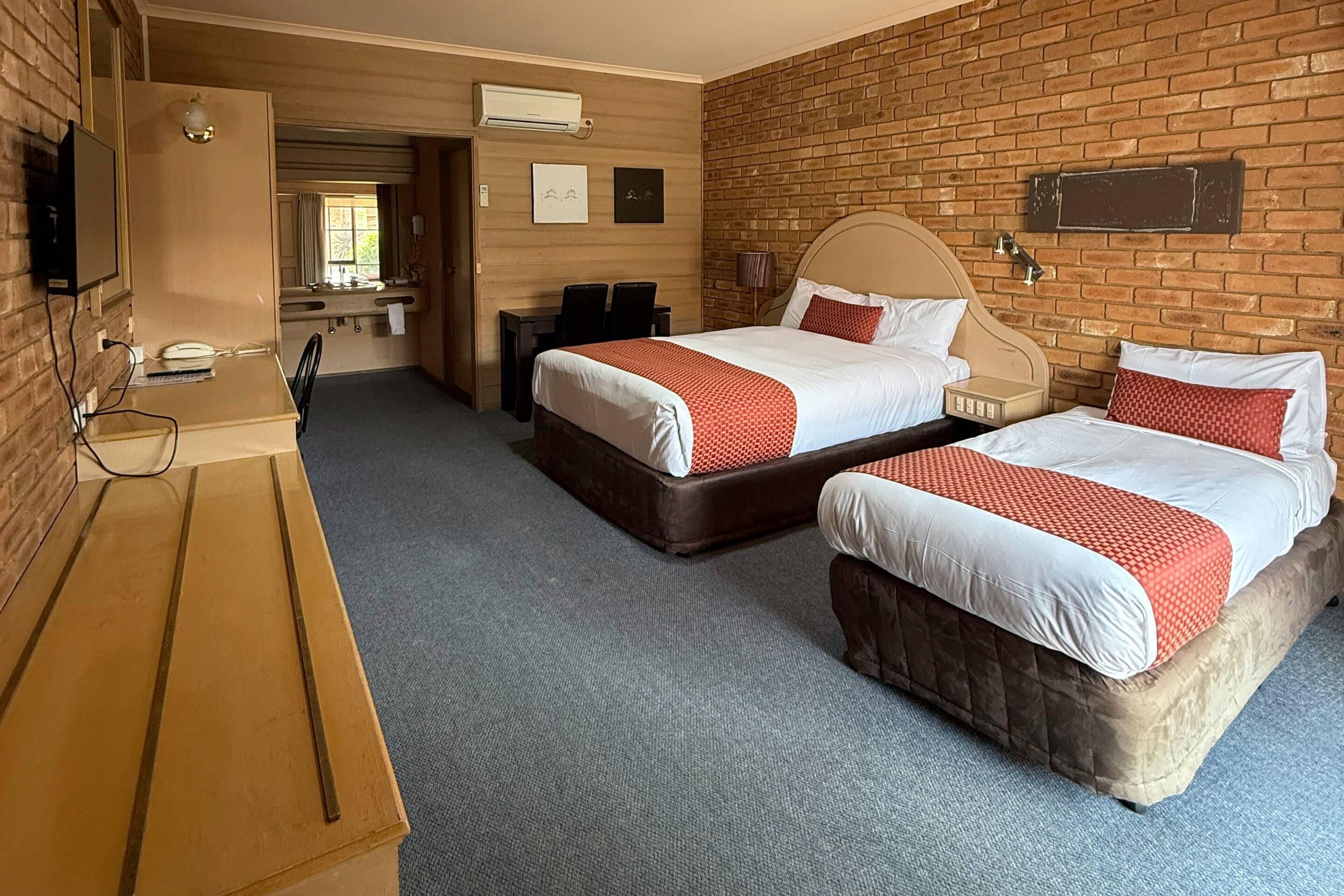 A hotel room with two beds, a desk, a TV, and a brick wall.