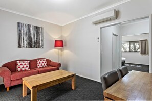 A room with a red sofa, a wooden coffee table, a dining table with chairs, a lamp, and a wall-mounted air conditioner. - Quality Inn Colonial (Golden Square)