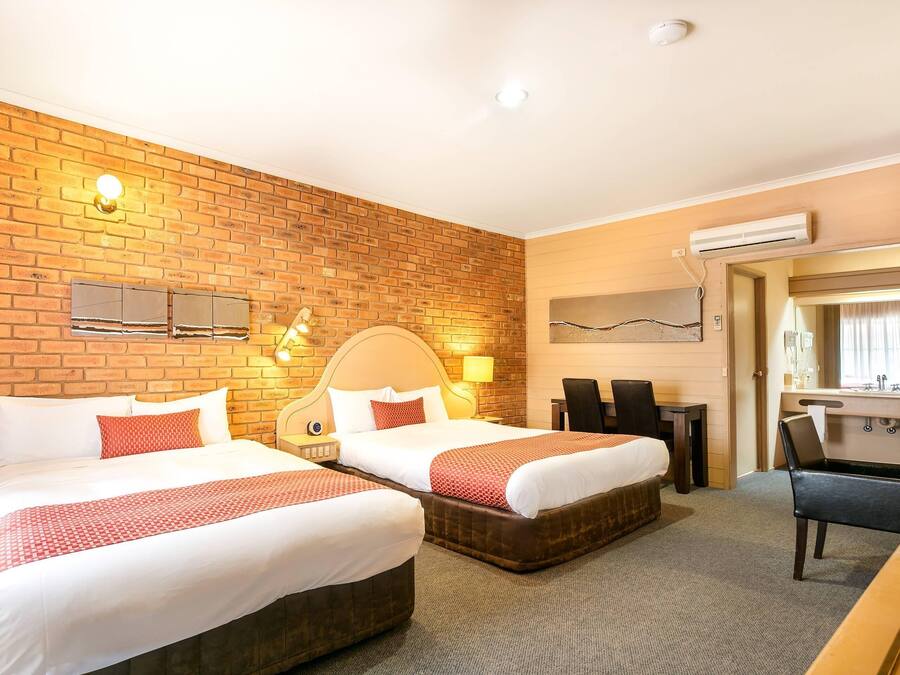 A hotel room with two beds, a brick wall, a wooden headboard, a desk, a chair, and an air conditioning unit.