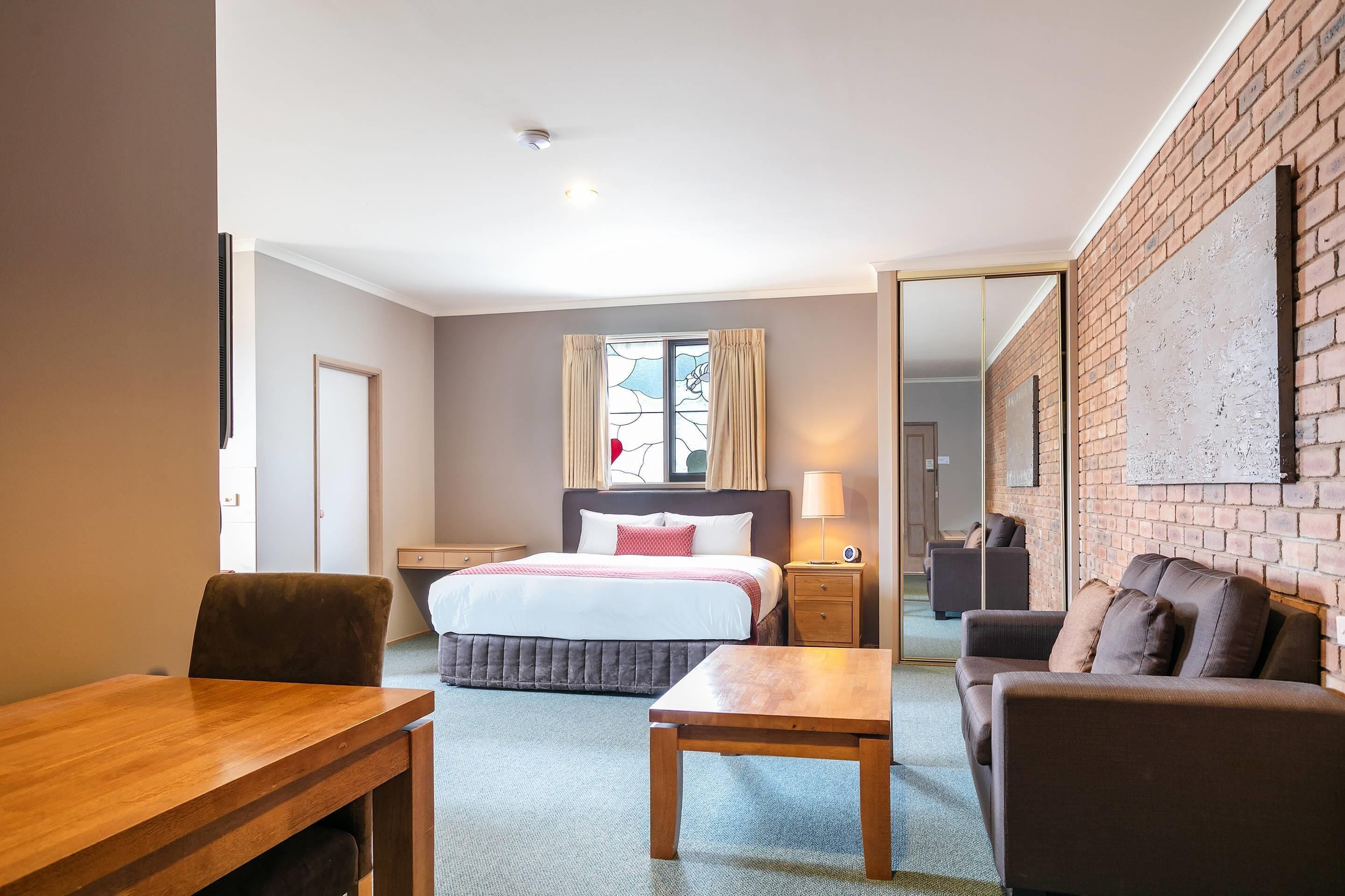 Une chambre d’hôtel avec un lit, une table en bois, un canapé, une table de chevet et une fenêtre avec des rideaux.