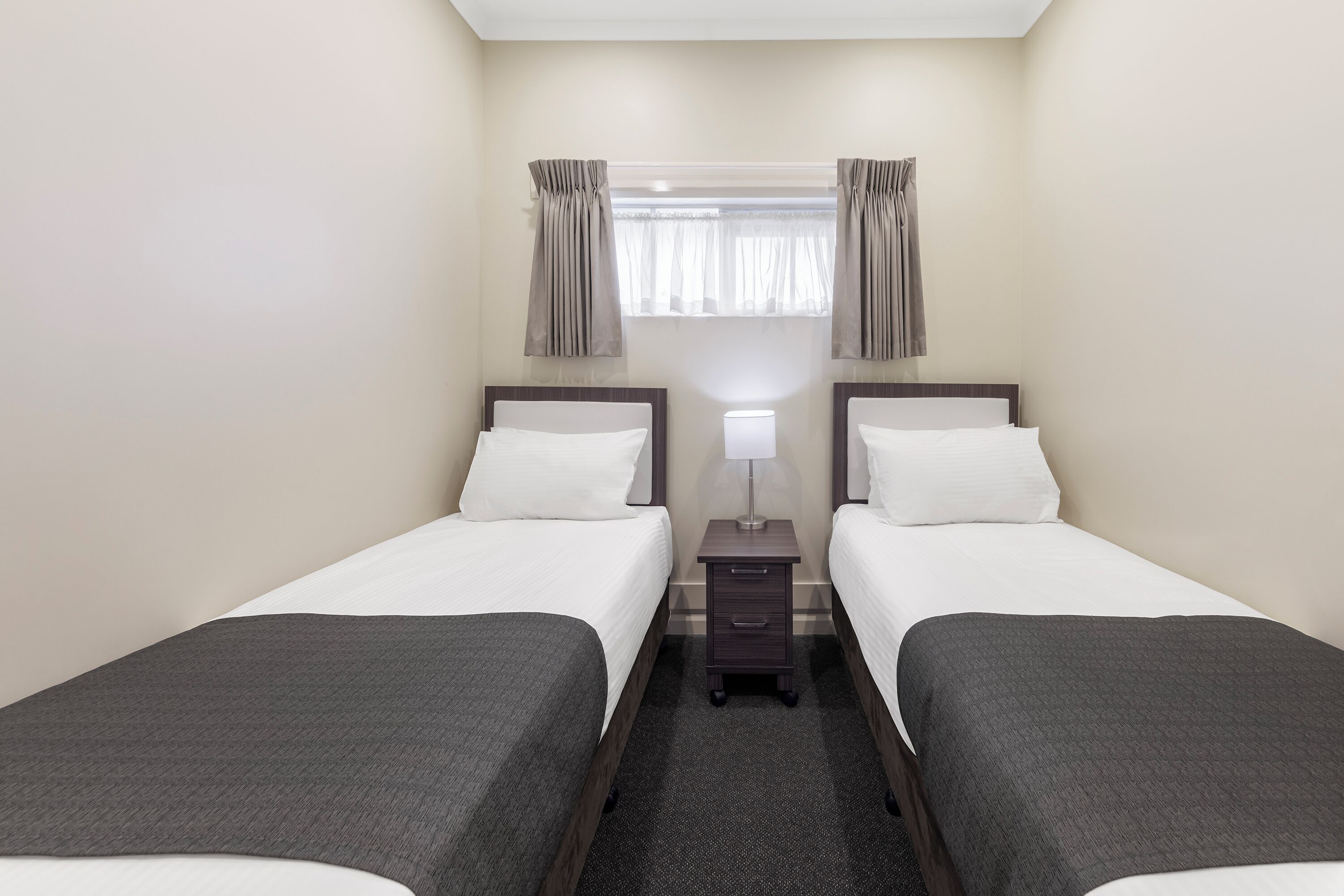 A hotel room with two single beds, a nightstand with a lamp, and a window with curtains.