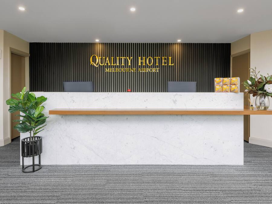 A hotel reception desk with a marble top, a black vertical panel with 'Quality Hotel Melbourne Airport' signage, two computer monitors, a small red object, and a plant on the left.