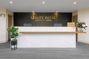 Ein Hotelrezeptionstresen mit Marmorplatte, eine schwarze vertikale Tafel mit der Beschriftung 'Quality Hotel Melbourne Airport', zwei Computermonitore, ein kleines rotes Objekt und eine Pflanze auf der linken Seite.