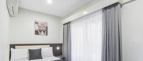 A hotel room with a bed, a nightstand, a wall-mounted air conditioner, and a window with curtains.