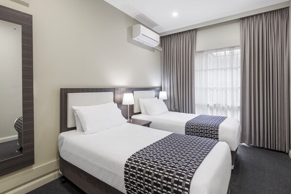 Habitación de hotel con dos camas, una mesita de noche, una lámpara, un equipo de aire acondicionado y una ventana con cortinas.