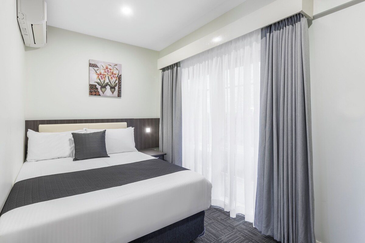 Habitación de hotel con cama, mesita de noche, aire acondicionado instalado en la pared y ventana con cortinas.