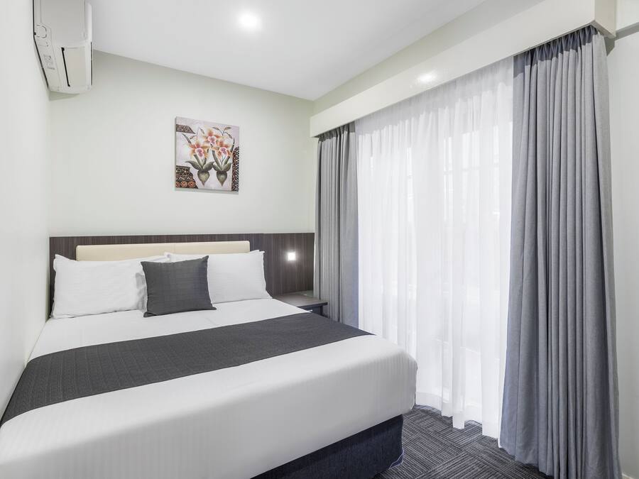 A hotel room with a bed, a nightstand, a wall-mounted air conditioner, and a window with curtains.