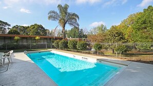 A rectangular swimming pool with a clear blue water surface, surrounded by a concrete deck. There are no diving boards, and a 'NO DIVING' sign is visible on the pool's edge. The pool is enclosed by a metal fence, and there are trees and shrubs in the background. - Moe Parklands Motel (Newborough)