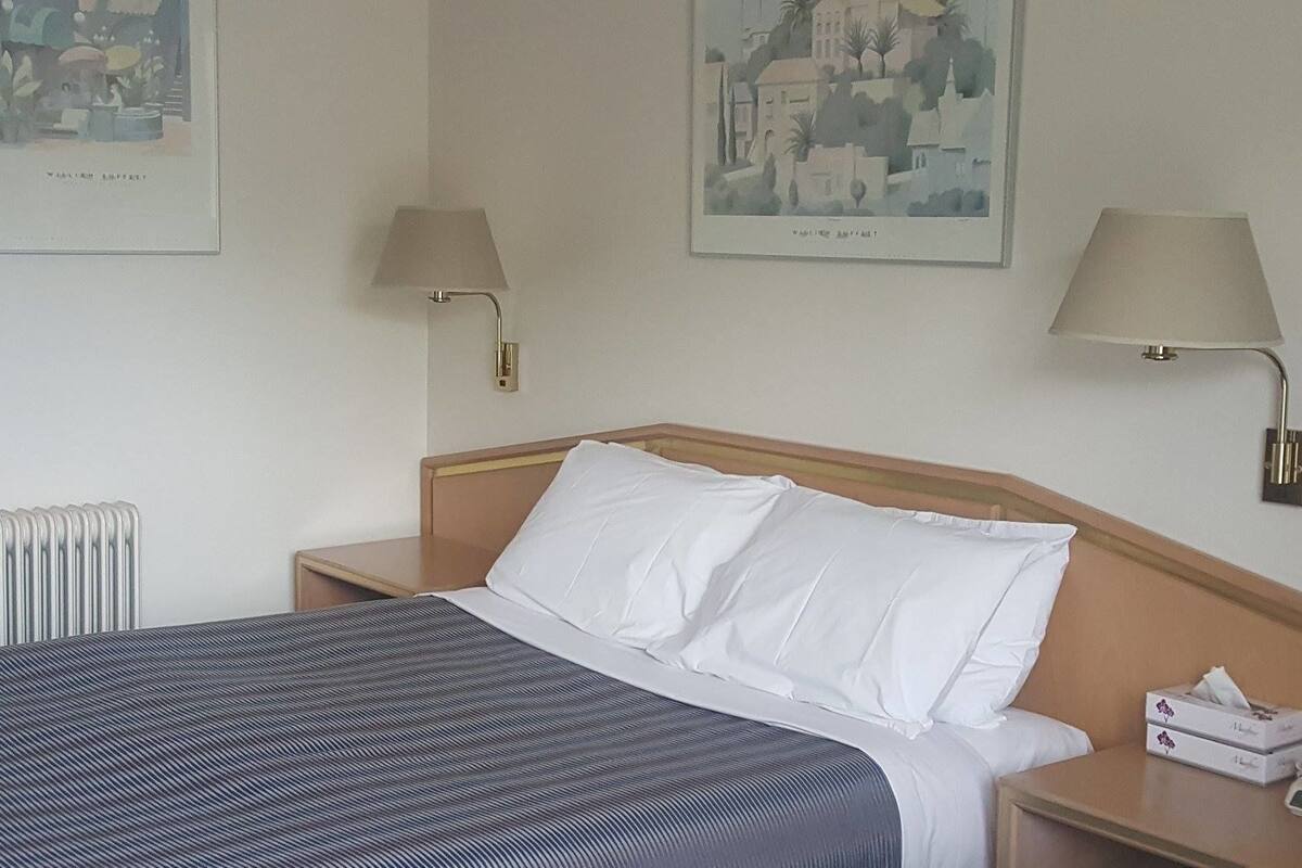Ein Hotelzimmer mit einem Bett, zwei Nachttischlampen, einem Nachttisch mit Taschentuchbox und zwei gerahmten Bildern an der Wand.