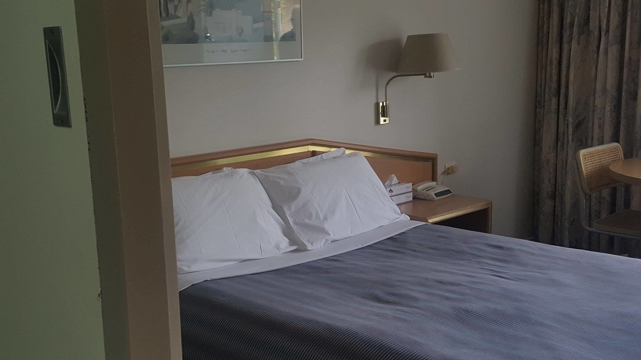 A hotel room with a bed, a bedside table with a lamp, a chair, and a wall-mounted telephone.
