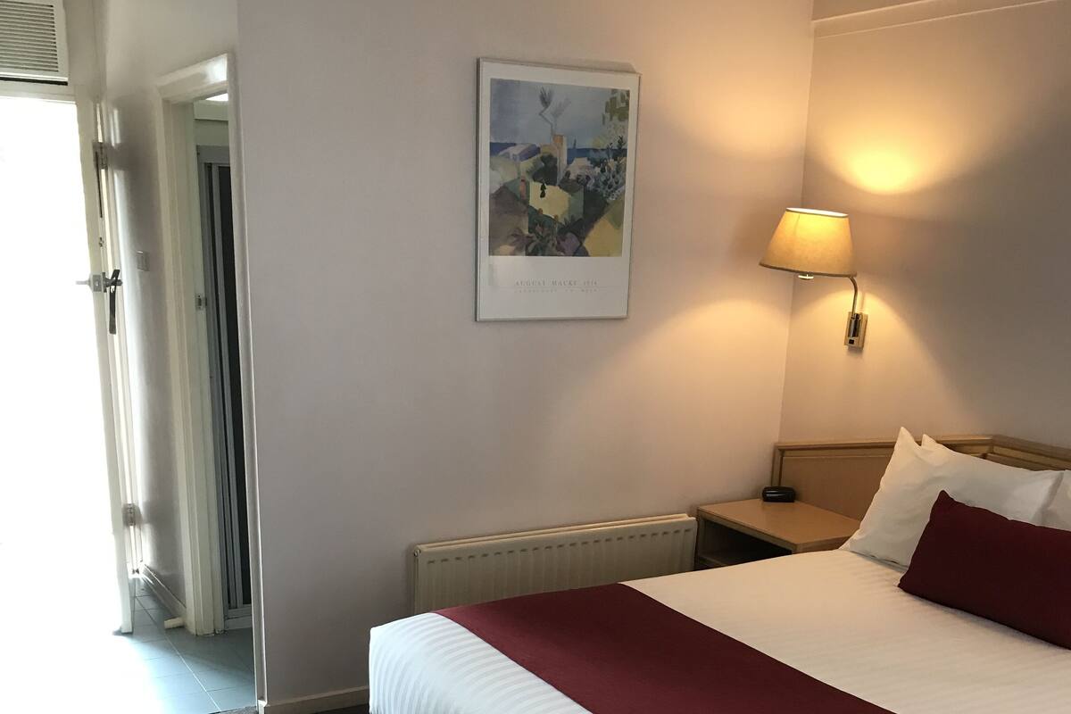 Ein Hotelzimmer mit einem Bett, einem Nachttisch, einer Lampe, einem Heizkörper, einem gerahmten Bild an der Wand und einer Tür zu einem anderen Raum.
