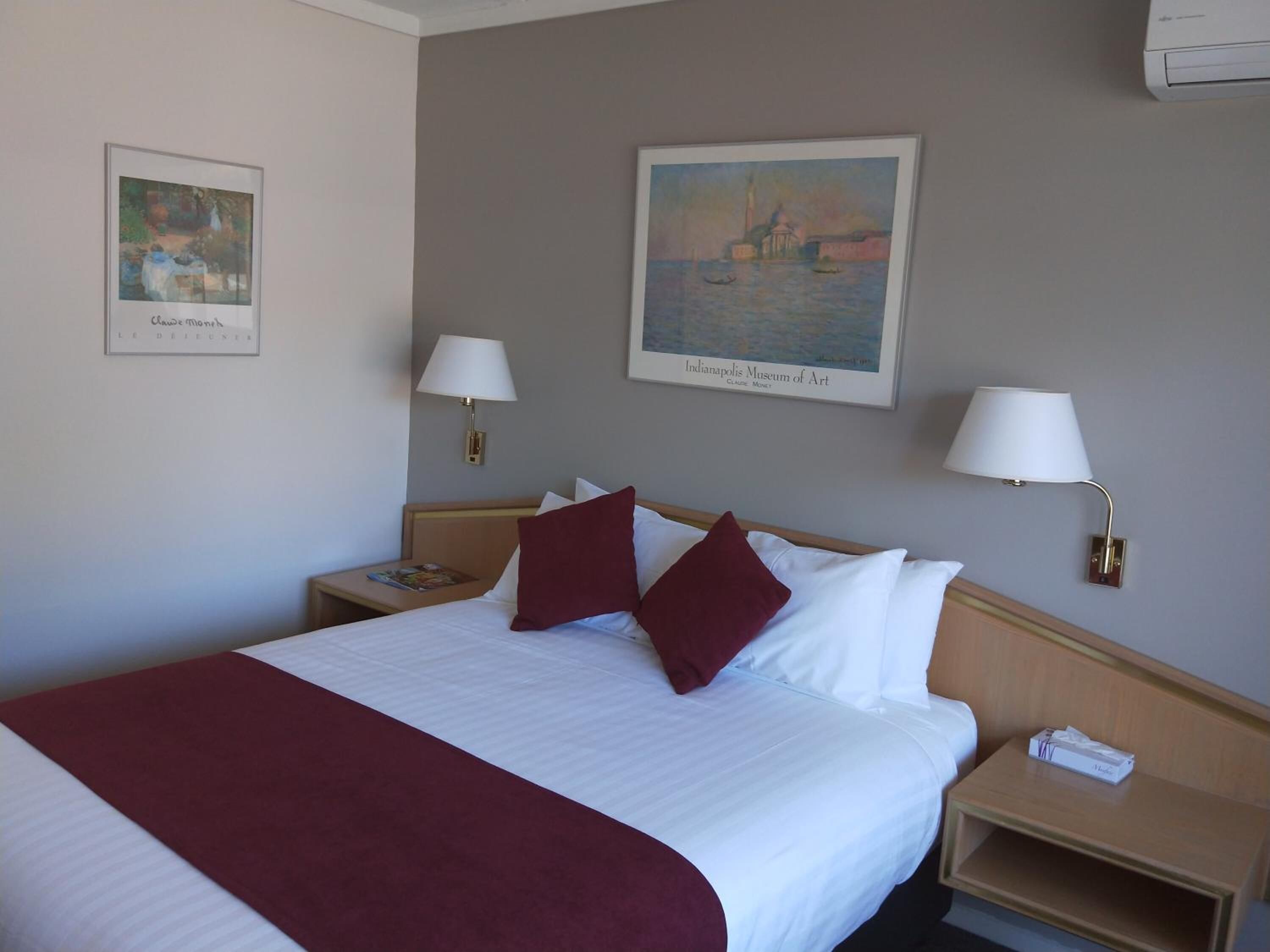A hotel room with a large bed, two bedside tables, a nightstand, and two wall-mounted lamps. There are two framed artworks on the walls.